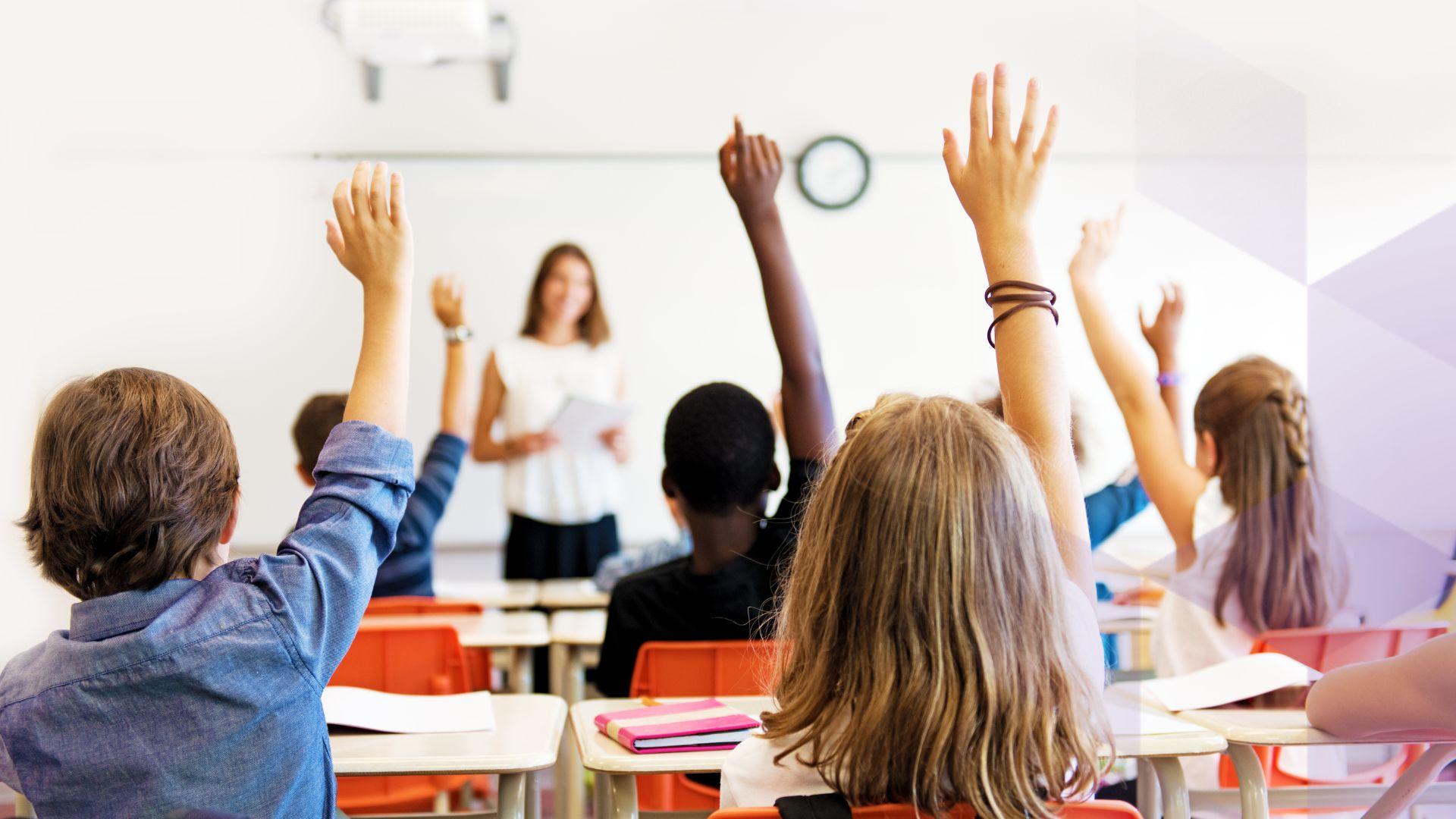 Children sit at desks with their hands up. A teacher stands at the front of the room
