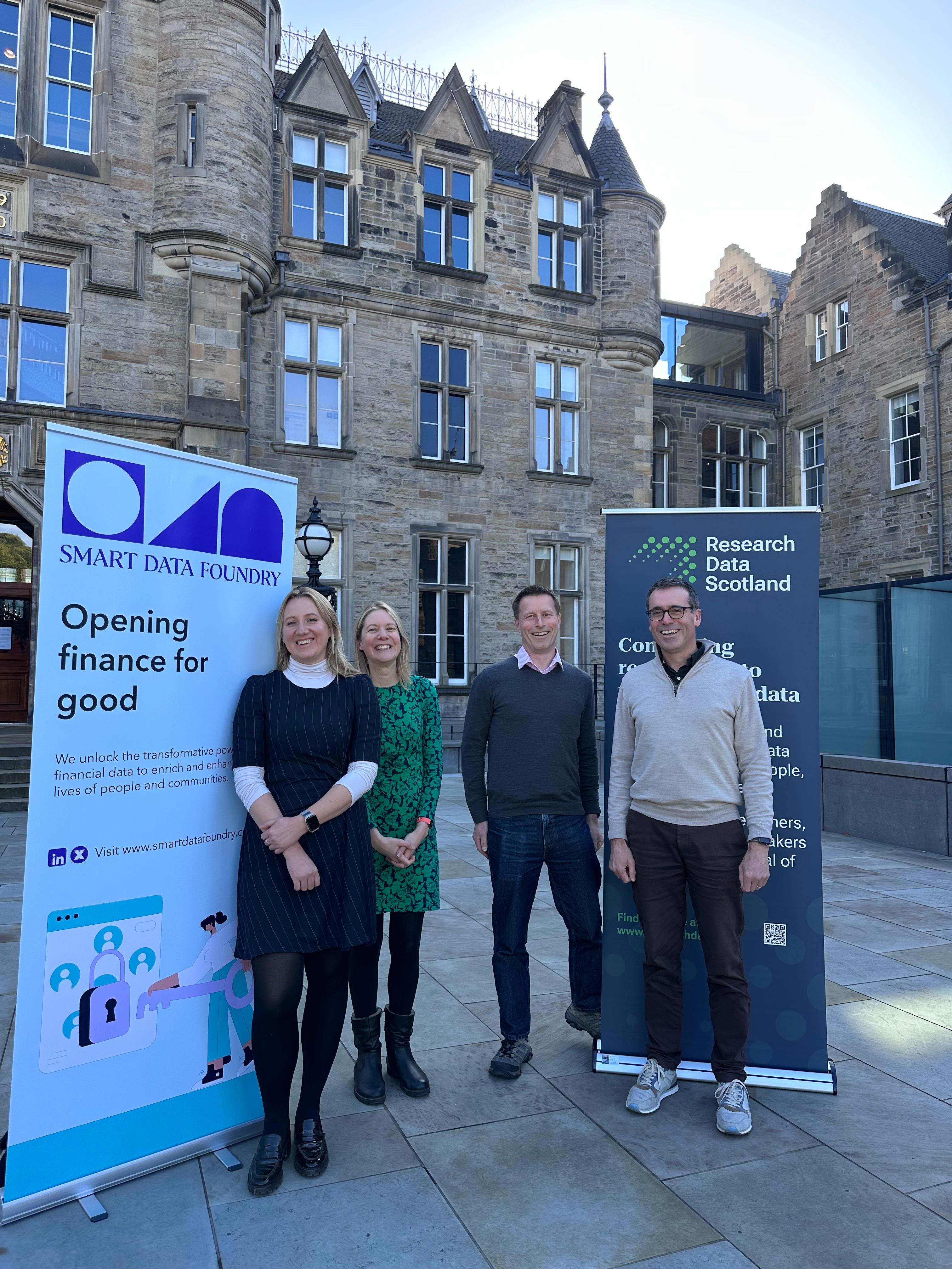 Four people stand smiling in front of an old building, with two pull up banner stands.